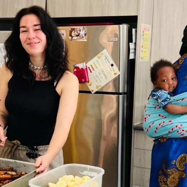 A photograph of two women in a kitchen preparing a large meal, while a baby is on the back of one.