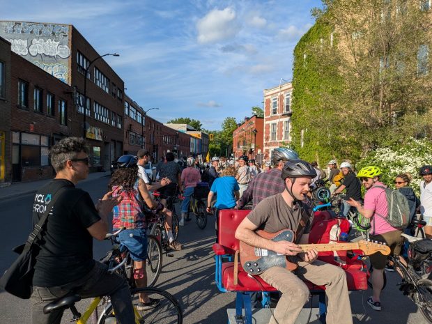 Critical mass of cyclists taking over a road. One of the bikes is pulling a car with seating, on which is a man playing an electric guitar.