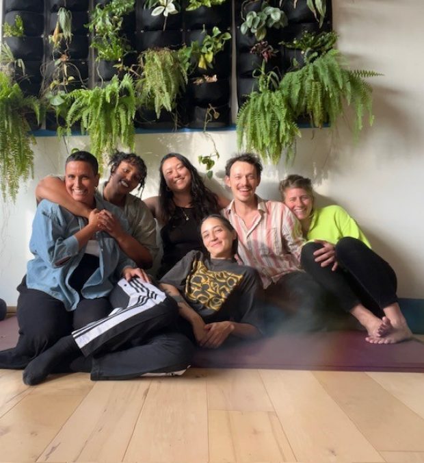 6 smiling people all huddled together on the floor. Behind and above them are lush green ferns.