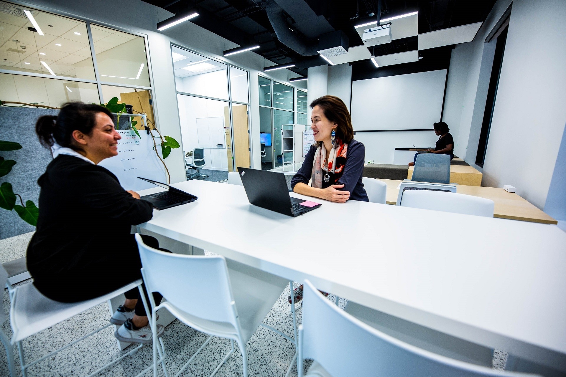 Two females are seated on highchairs and table in the co-working space, smiling at each other, another one is seen further in the back seated on a block. 