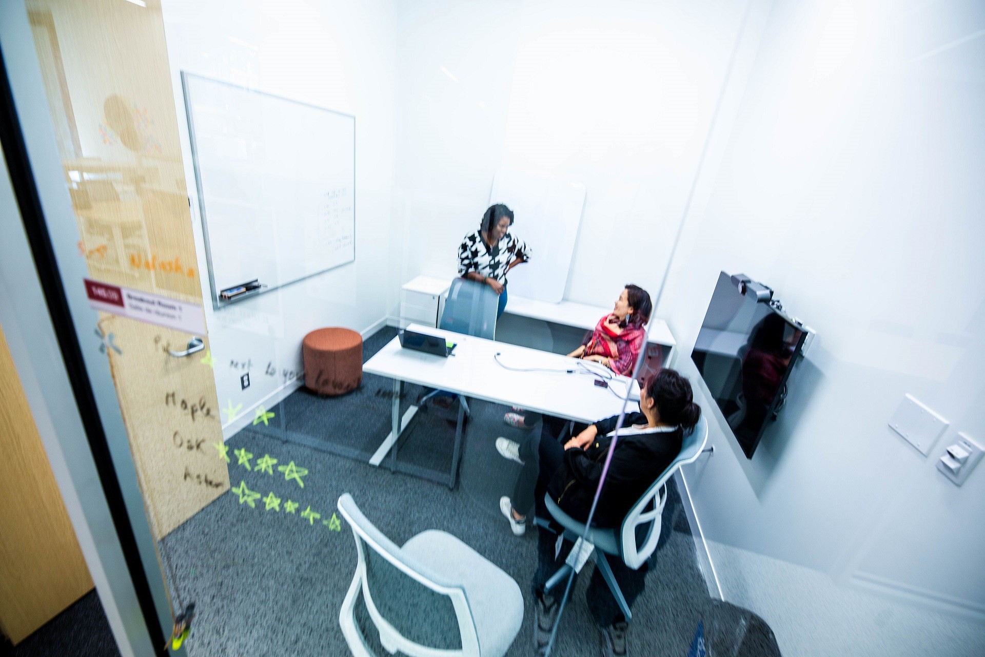 Three females, 1 standing, two seated, are discussing in a room with clear glass walls, a white table, a white board and a TV screen. 