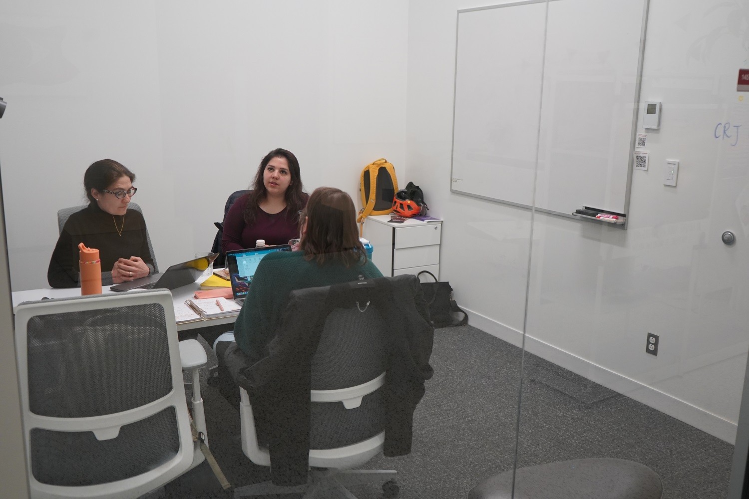 3 peoplesitting in the breakout room and working on their laptop. The room has clear glass walls and includes a white table and a white board