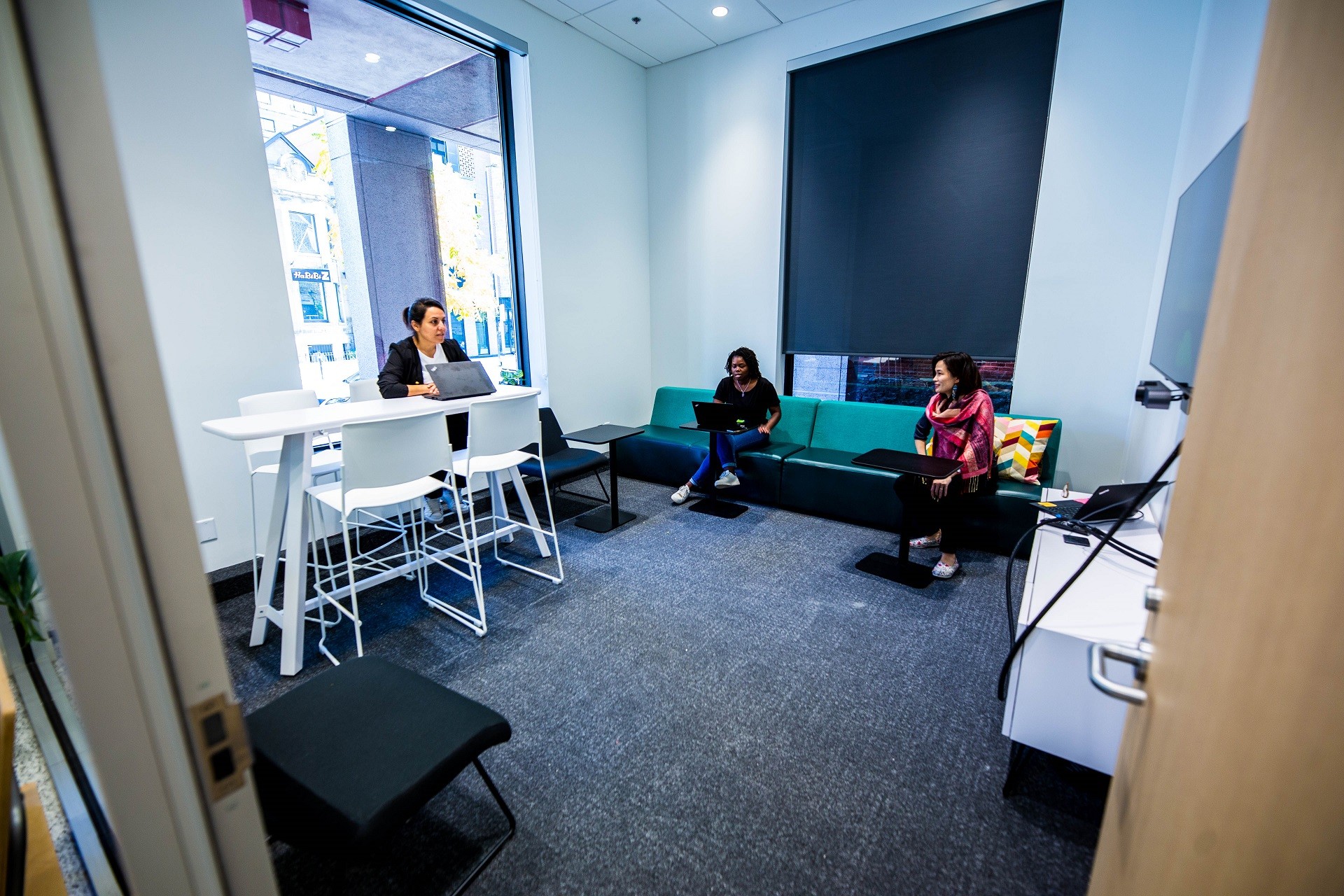 Three females are working in the lounge room: 1 is seated on a high white chair with their laptop on a high white table; the two others are seating on a long green couch. The room faces the street side, visible through the large glass window. 