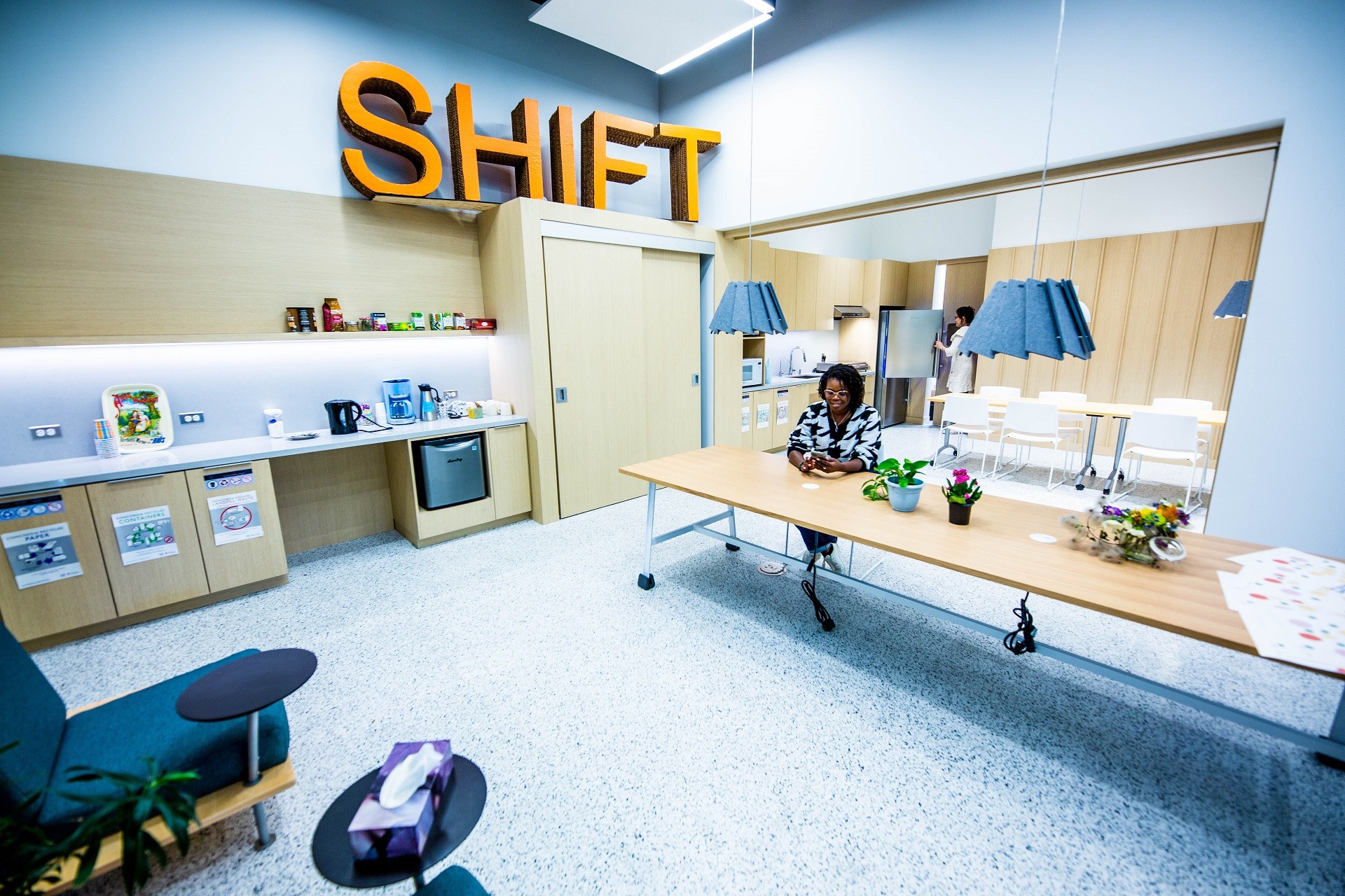 A female is sitting in the dining area looking at her phone, the area includes two long tables, a counter with coffee and tea maker and large SHIFT letters at the top of the shelves. 