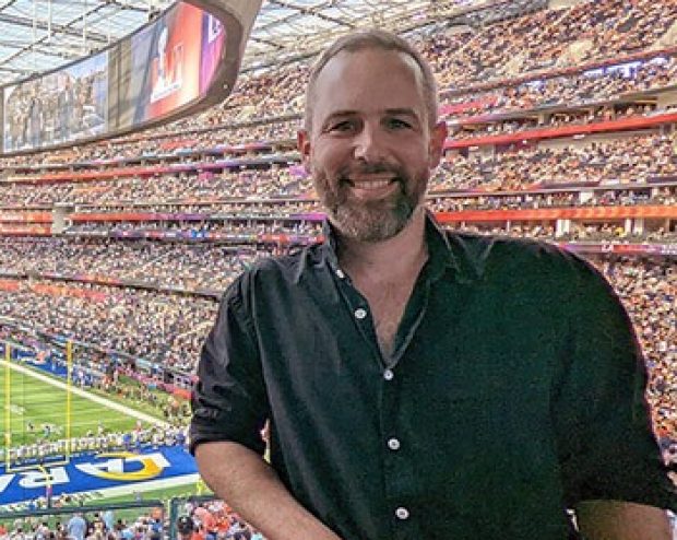 A man with short hair and a beard, wearing a black shirt, standing in a crowded stadium with a smile.
