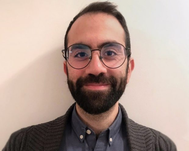 A man with a beard and glasses, wearing a grey sweater over a collared shirt, smiling in front of a plain white wall.