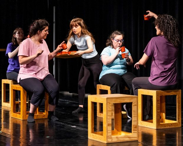 Five individuals sit on wooden seats on a stage, acting out a scene