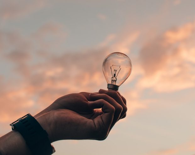 picture of a hand holding up a light bulb, with sky in the background