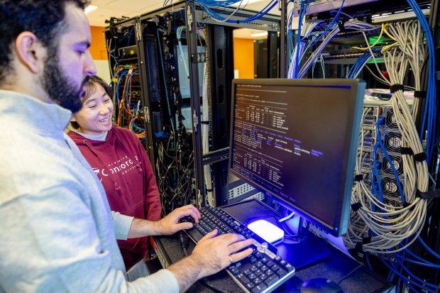A man types on a keyboard with monitor and server in the background as a woman looks on. 
