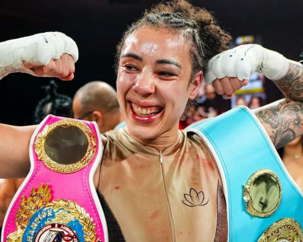 A boxer in the ring flexes both arms while wearing two championship belts, one pink and one light blue, draped over the shoulders after a match.
