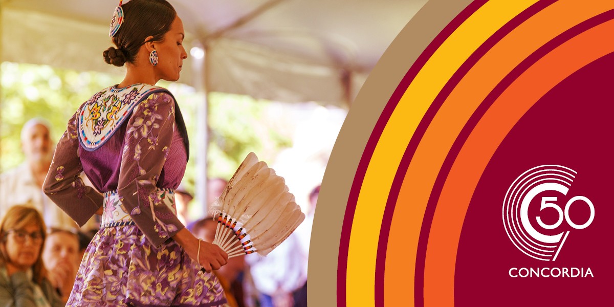A dancer at a Pow Wow stands with her back to us, wearing a brightly coloured shawl i