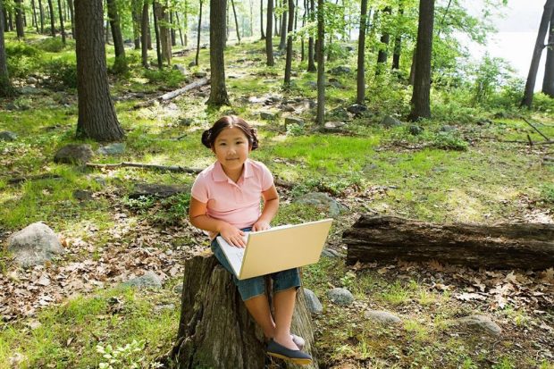 A young girl wearing a pink polo t-shirt and cropped jeans is sitting on a stump in the woods with a laptop on her lap.