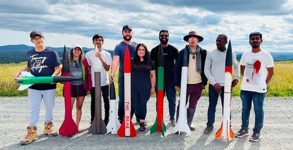 Nine members of the team are standing in a row outside holding up models of their rockets