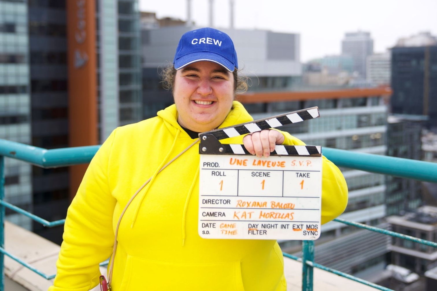 Roxana Biloiu wears a yellow sweatshirt and a blue cap with the word CREW on it and holds the clapperboard for her film "Une laveuse svp"