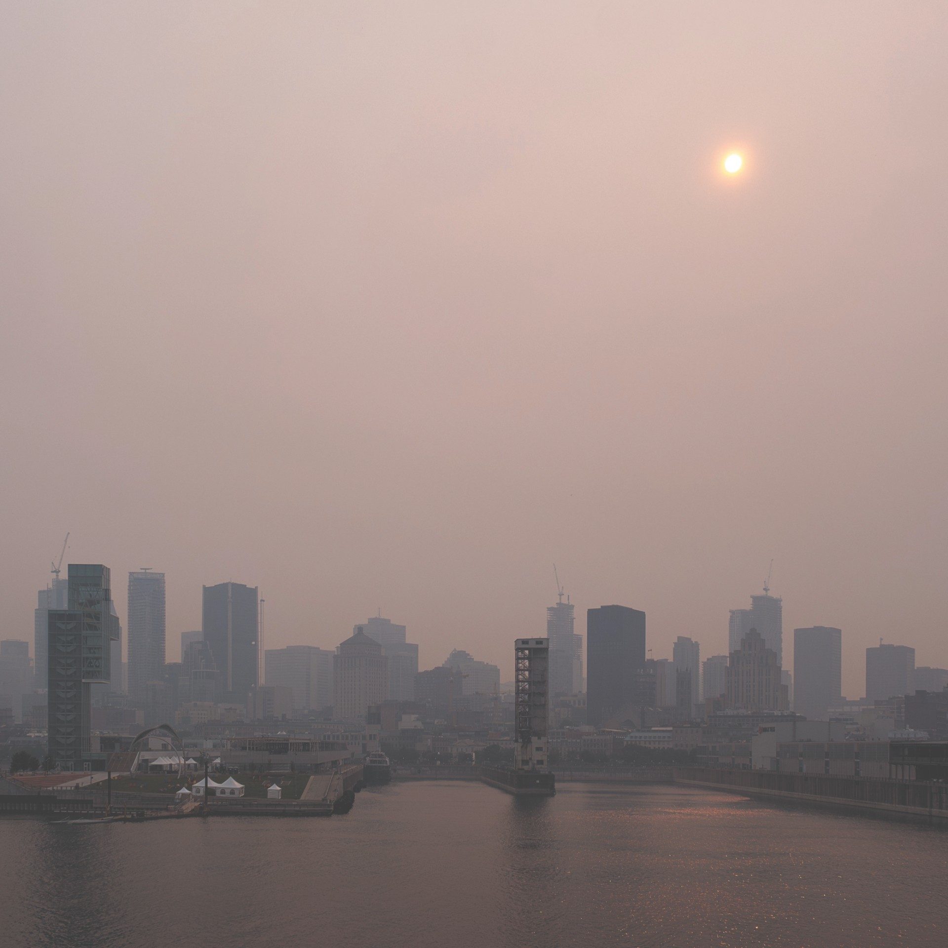 A hazy skyline of Old Montreal with muted colors, featuring the sun as a pale disc in a smoggy sky. The Saint Lawrence River is in the foreground reflecting the city's silhouette, and various buildings are visible, some under construction. There's a sense of calm despite the overcast atmosphere, with a small boat visible on the water.