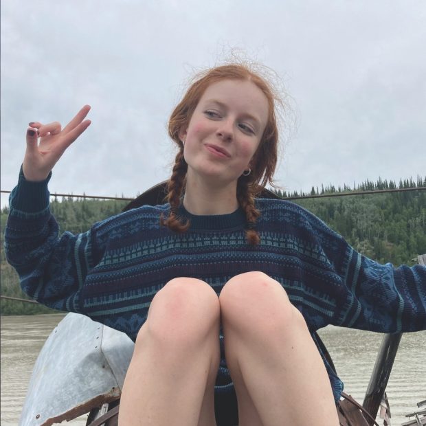 A woman with red hair styled in braids is sitting and smiling at the camera, making a peace sign with her fingers. She is wearing a blue and black patterned sweater and is seated in front of a calm river with a dense forest in the background. The sky is overcast.