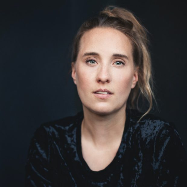 Photo of a woman with a dark tshirt and blonde hair in a ponytail looks into the camera in front of a dark wall.