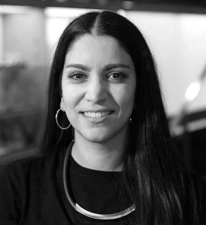  The image is a black and white portrait of Marta Samuel, the Editor of the Concordia University Magazine. She has a warm smile and is looking directly at the camera. Marta has long, straight hair, and she is wearing a dark top with a crew neckline and a distinctive, light-colored necklace. Her expression is friendly and approachable.