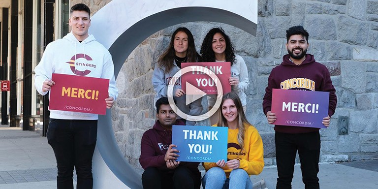 Six students holding signs that say "Thank You" and "Merci" stand next to the Concordia big "C" in front of the Hall Building