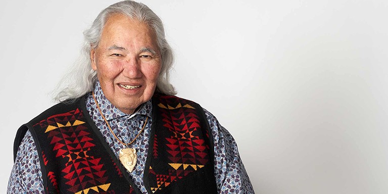 A man with long grey hair and a vest sits and smiles.