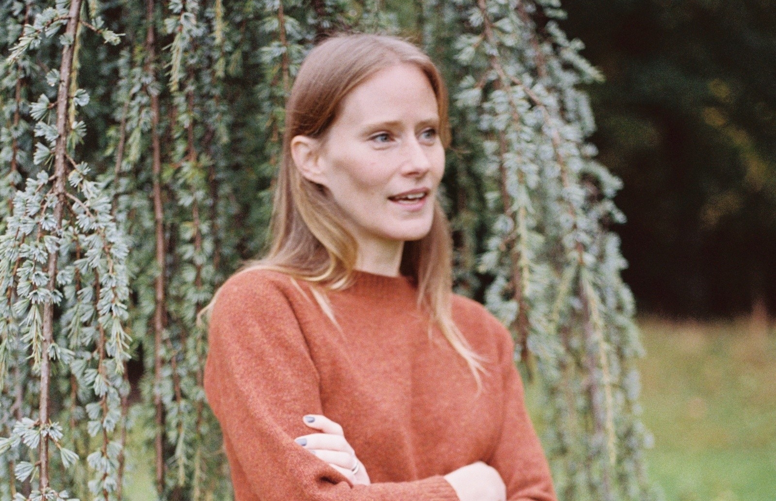 A woman with long blonde hair and a red sweater stands outside in front of a tree