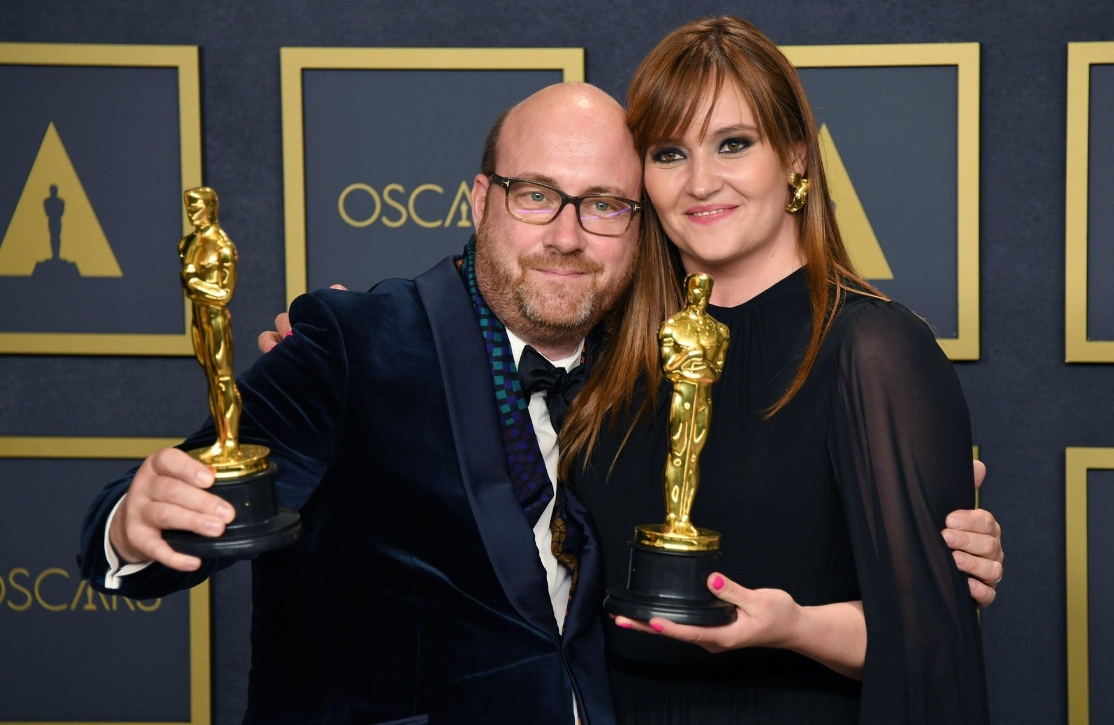A man and a woman pose for a photo. They are dressed up and are each holding an Oscar Award in their hand.