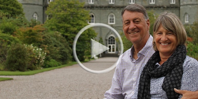 Constantine Constandis and Anne Upton-Constandis sit on a white iron bench on a path in front of a Scottish castle