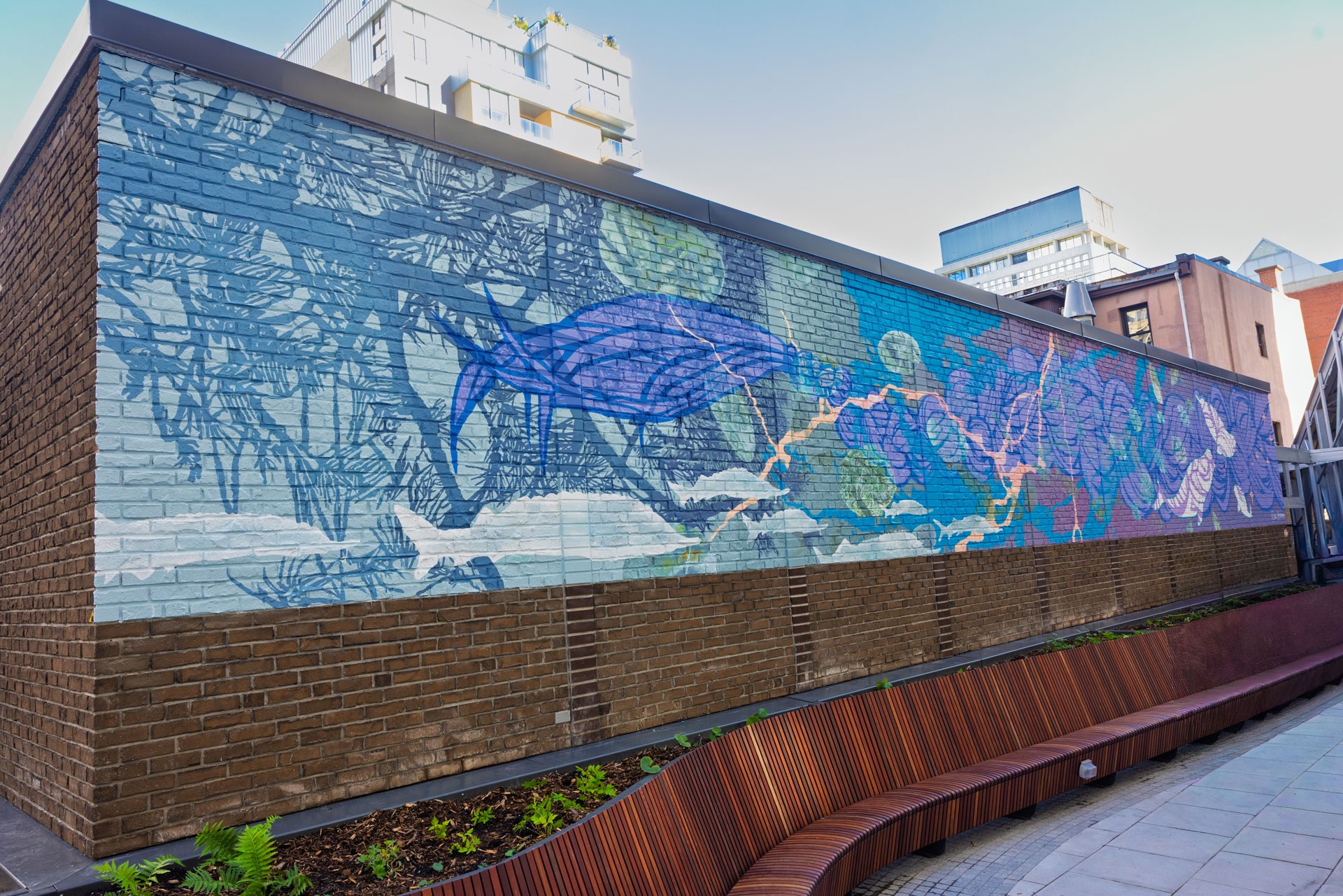 Une murale sur un mur de briques représente le pin blanc, l'esturgeon, la rivière, des plantes médicinales, une tresse, la constellation des Pléiades et des bélugas dans des nuances de bleu, d'aigue-marine et de violet.