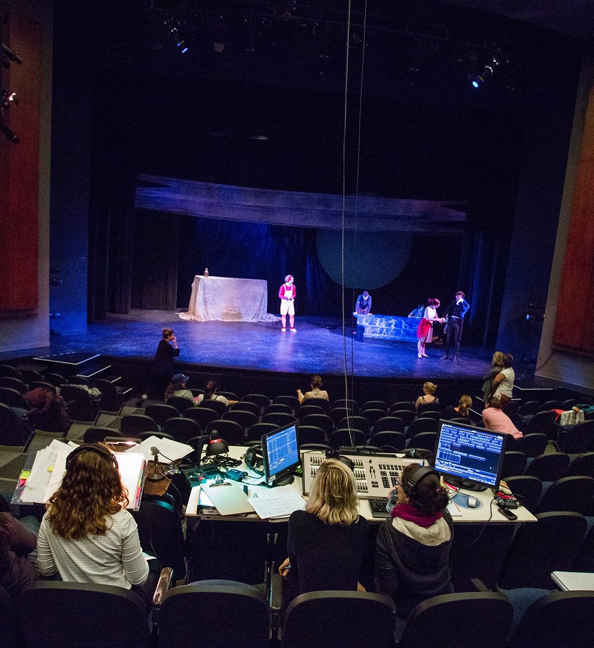 Technical rehearsal in DB Clarke theatre Auditorium featuring few people on stage, few people sitting in the theatre, and a technical crew consulting papers and computer monitors