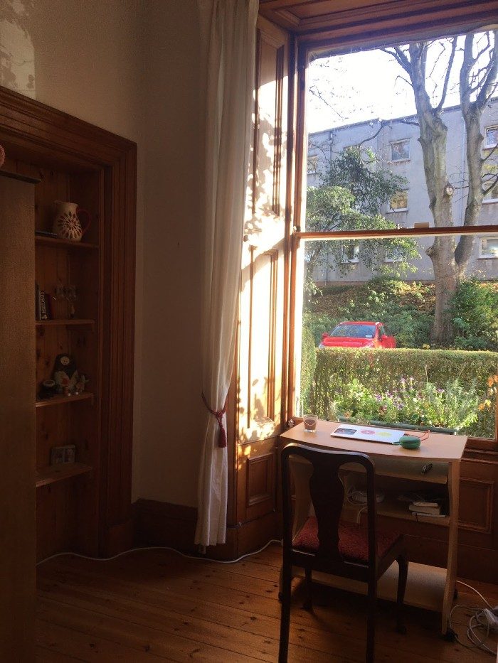 A small desk and a chair face a large sunny window in Ceilidh Michelle's space