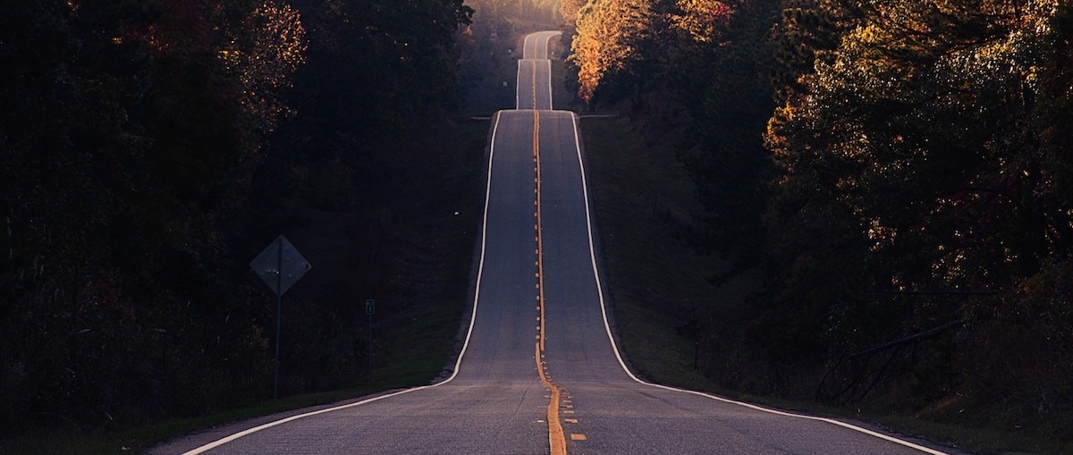 pictures a pavement road rising and falling into the distance with a sunset on a forest in the background