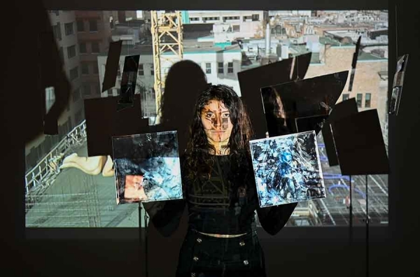A photo of Sabina Gámez holding two paintings against a cityscape, with the cityscape also projected across her face