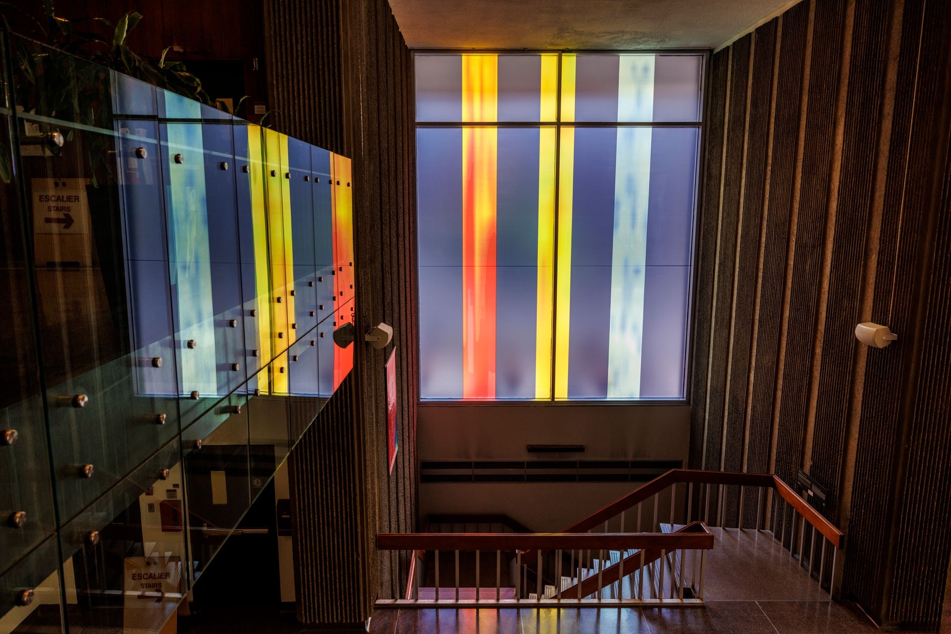 Interior view of a stairwell in a building. Tall, vertical translucent panels in bright colors (yellow, red, blue and orange) fill a window at the top of the stairs, casting colored light and reflections into the space. 