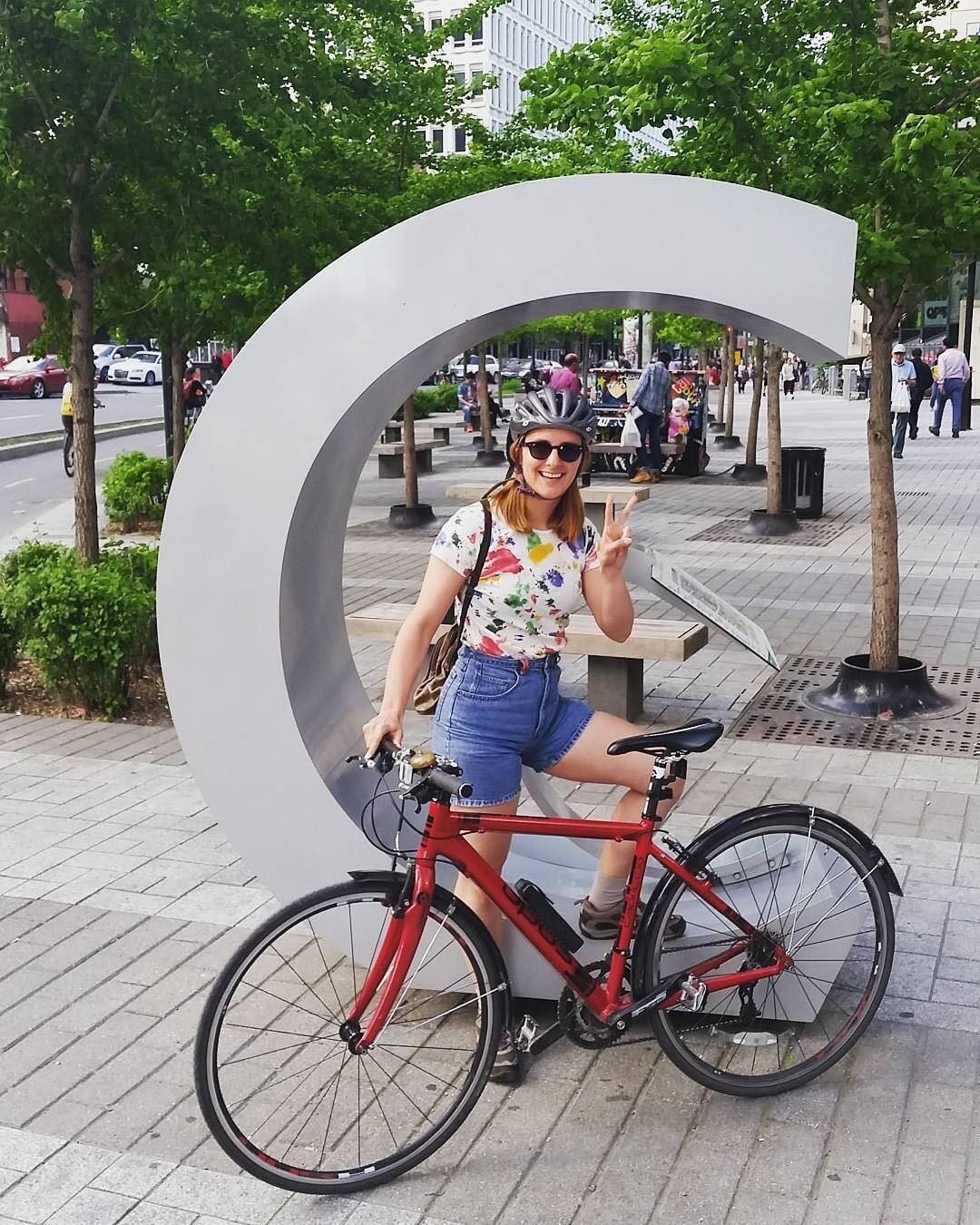 A smiling young woman poses with her bike in front of Concordia's letter C logo sculpture
