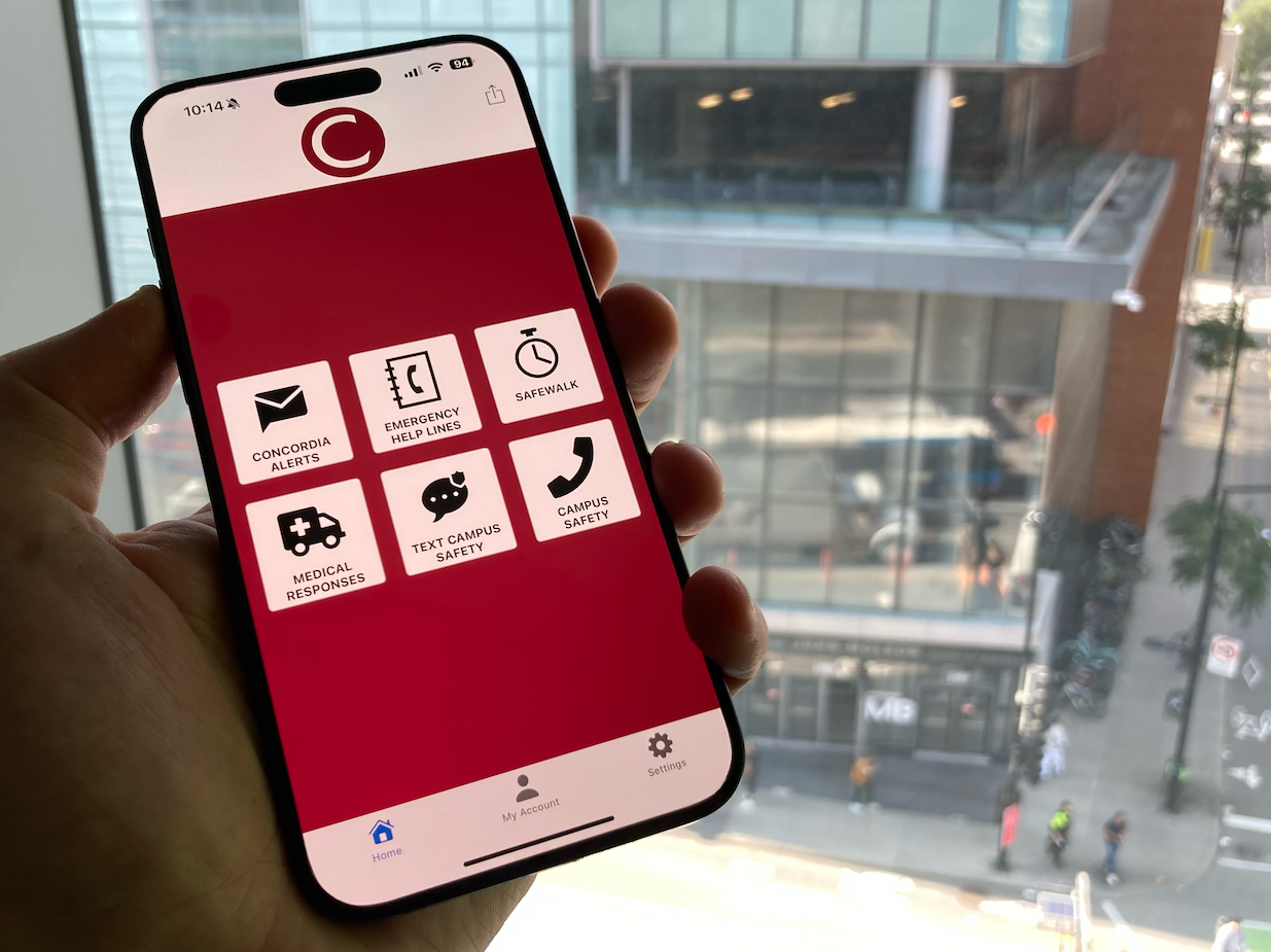 Person holding a smartphone showing the Concordia safety app with options for alerts, emergency help lines, Safewalk, medical responses, texting campus safety, and campus safety information. A campus building is visible through the window in the background.