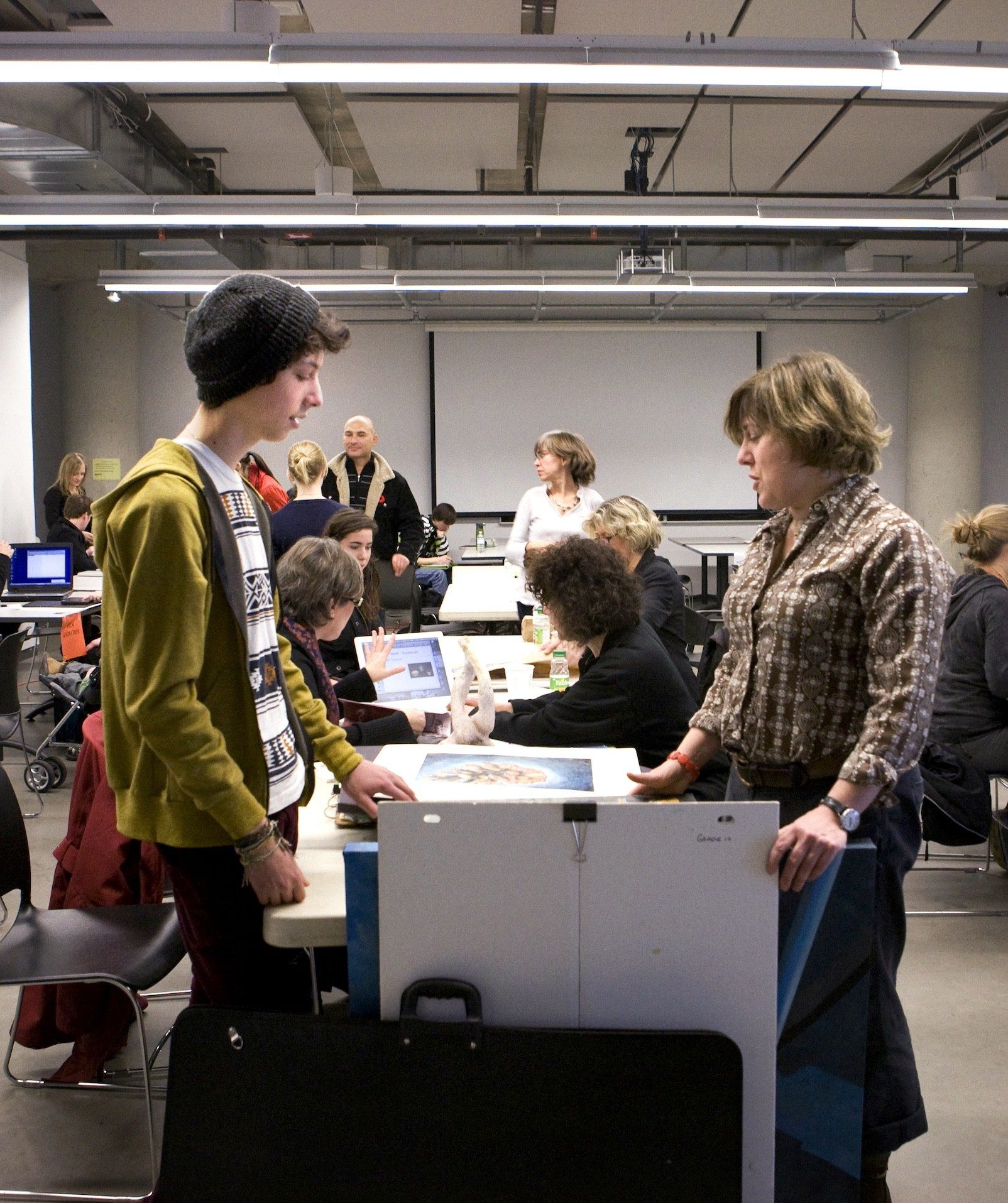 Male student and female instructor standing across from each other looking down at art work on table. Male student and female instructor standing across from each other looking down at art work on table.