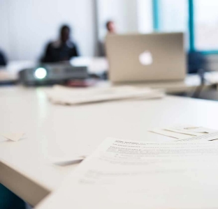 A circular blurred photo of printed text sheets on a white desk in the foreground with the back of a laptop computer and two seated people in the background. A circular blurred photo of printed text sheets on a white desk in the foreground with the back of a laptop computer and two seated people in the background.