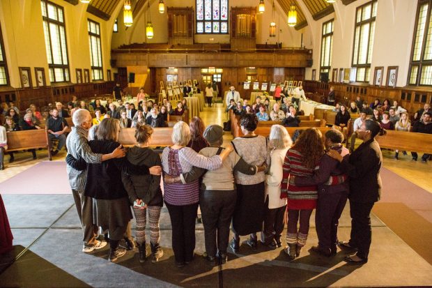 ten faculty standing side-by-side with arms around each others shoulders in front of a large audience in a hall for an awards gala