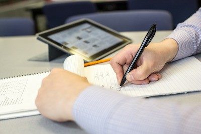 A person is writing in a notebook and there is a tablet on the table