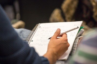 A person is writing in a notebook that is being steadied on his calf as he sits with one leg crossed over the other in a chair.