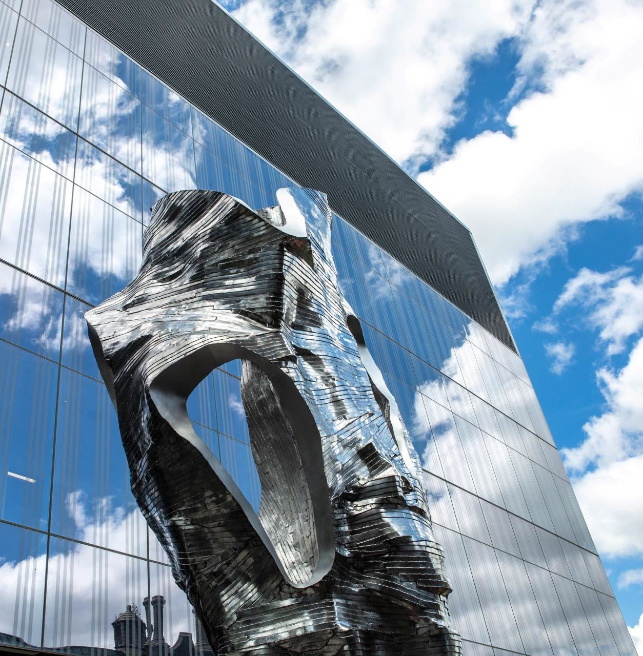 Metallic sculpture outdoor in front of tall building reflection blue sky and clouds.      
