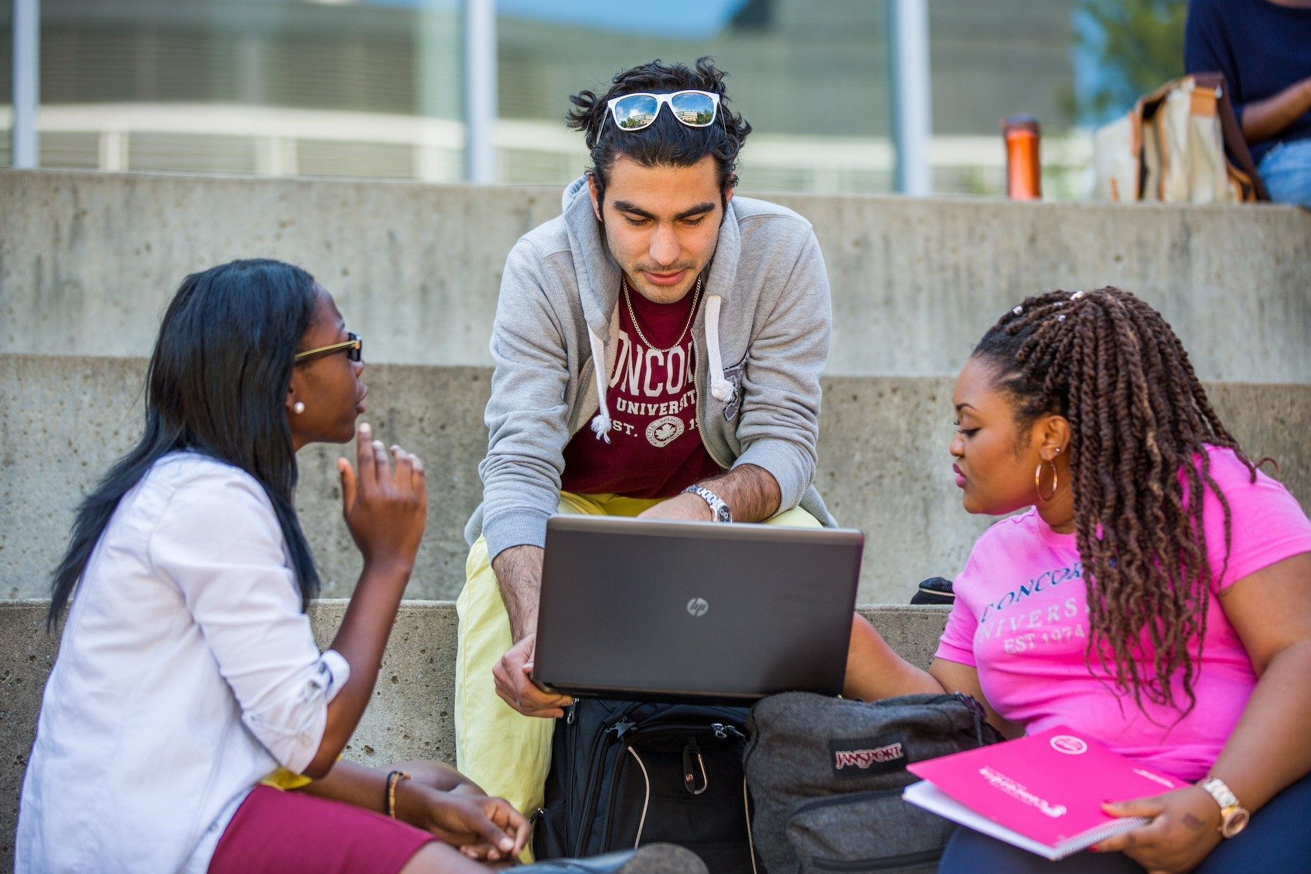 Two female students and a male student sitting together outdoors on steps working around a laptop.