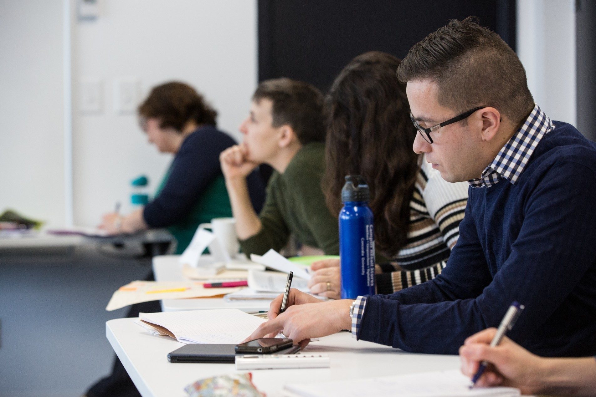 A male student sitting in class taking notes with a few other surrounded by a few other students.