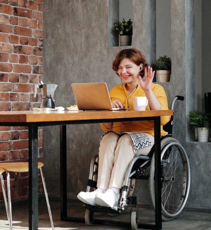 Woman in wheelchair waving hello in front of a laptop and wearing earbuds