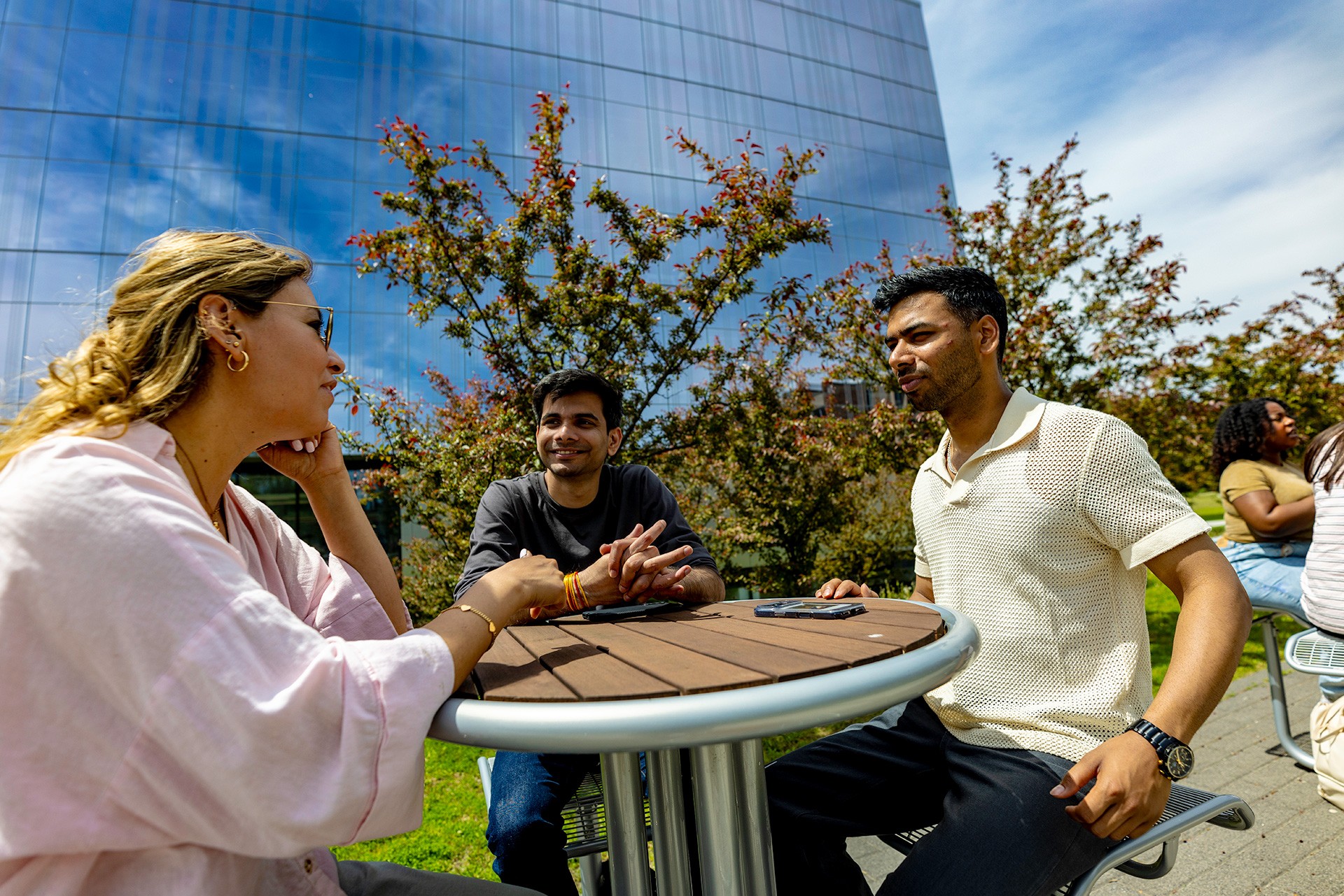Three students, one woman and two men sitting outside at a round table talking.