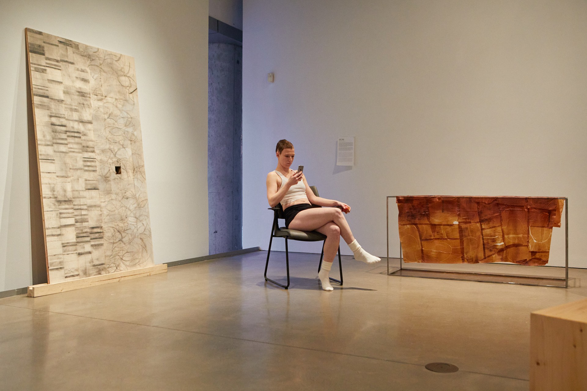 Anne-Marie Latulippe (a.k.a “Steel Toes”) is documented sitting next to  on a chair, holding a phone in their left hand and an apple in the right. She is filming the audience as they are circulating in the gallery space.