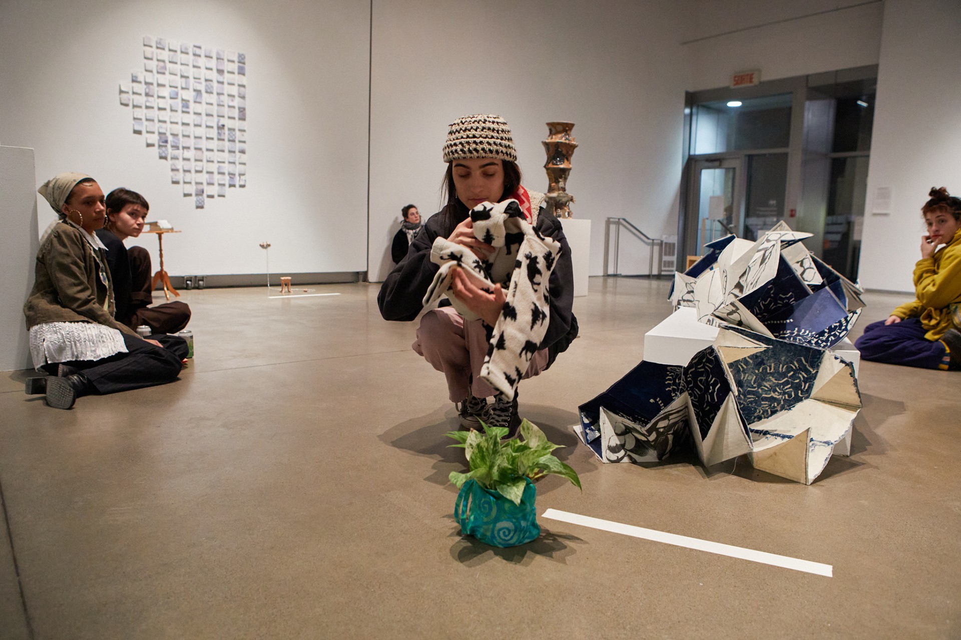 Laura, the performer is captured squatting in the middle of the gallery while holding different objects given by friends, in this scene we see most predominantly a black and white sweater and a house plant. 