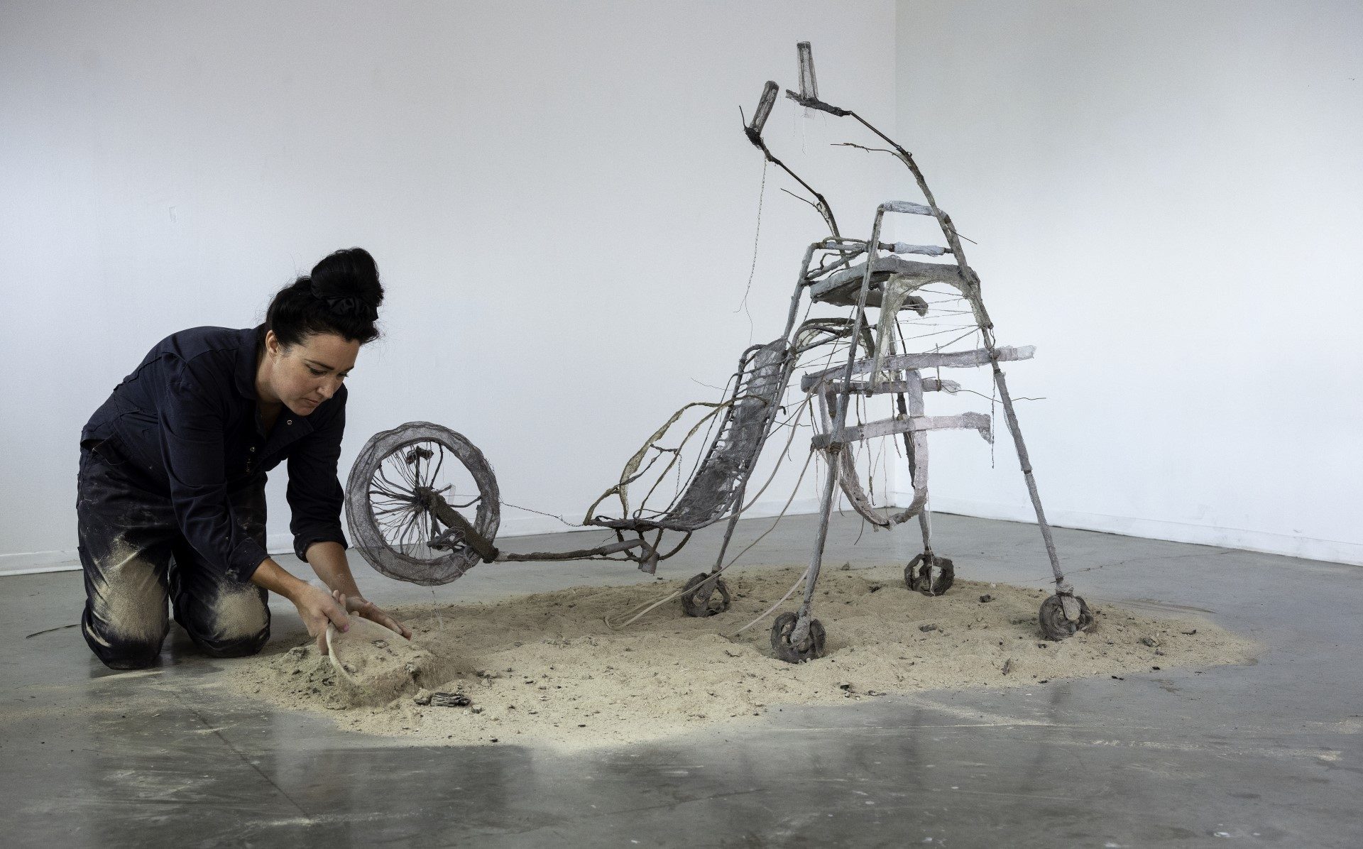 Female artist kneeling in a pile of sand next to an abstract sculpture