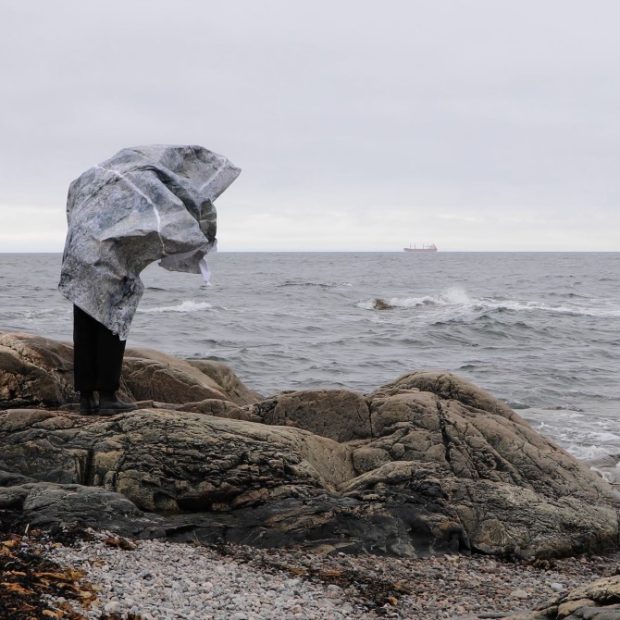 Woman artist standing on rock in front of water covered with a large paper overlaid with a rock pattern