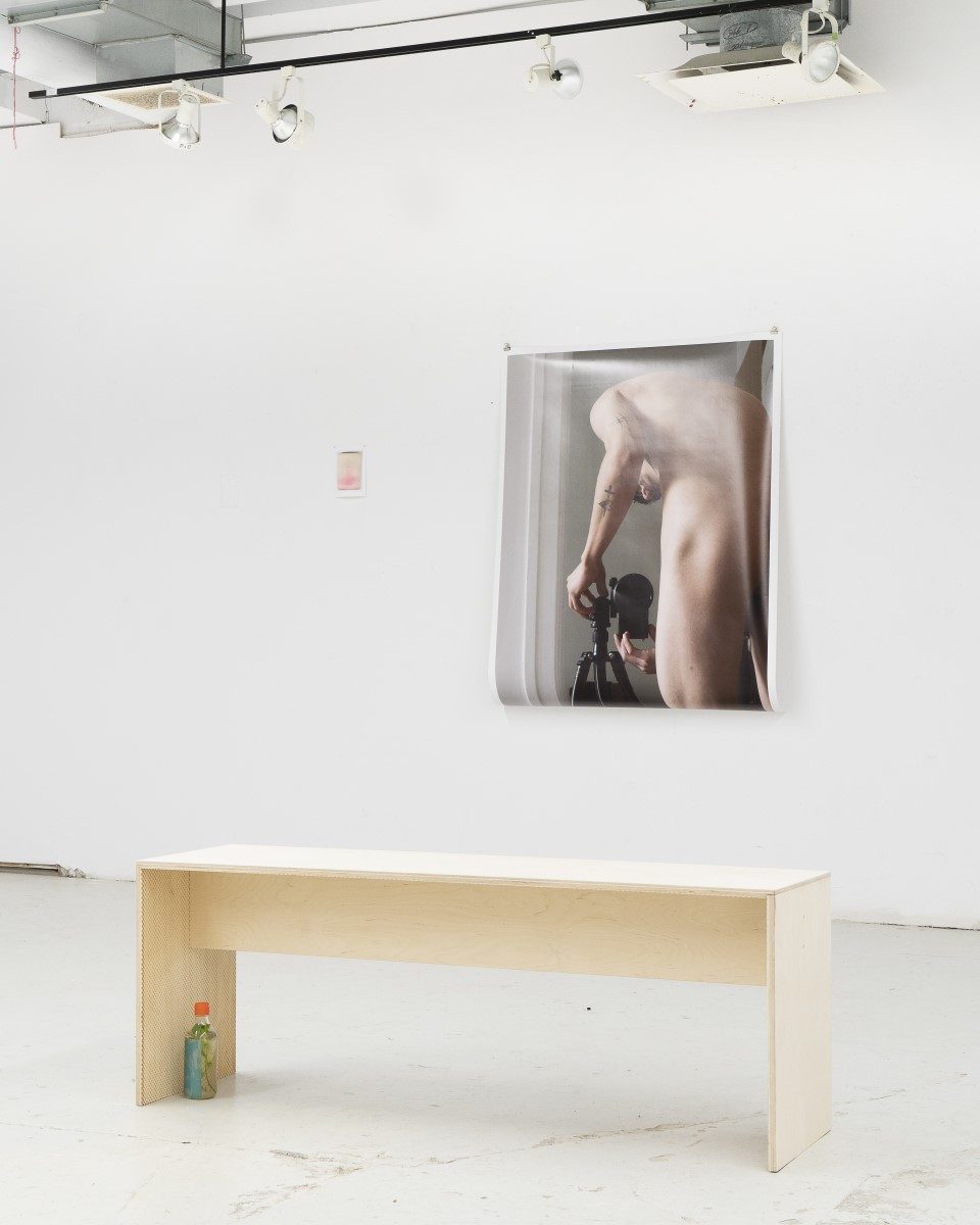 Installation view of a past exhibition featuring a nude self-portrait of the artist and a hand-made wooden bench with engraving.